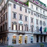 A historic beige, multi-story building with arched windows and balconies, adorned with flowers. Several flags, including Italys, hang by the entrance, and ground-floor shops face a quiet street.